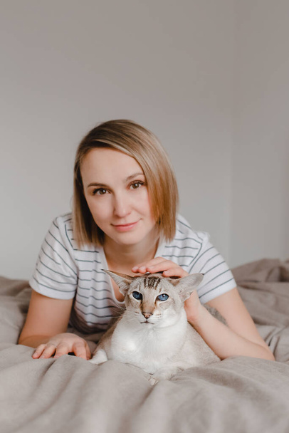 Proud pet owner. Happy Caucasian smiling blonde woman petting oriental cat with blue eyes. Beautiful girl lying on bed in bedroom at home and stroking domestic feline animal.  - Фото, изображение