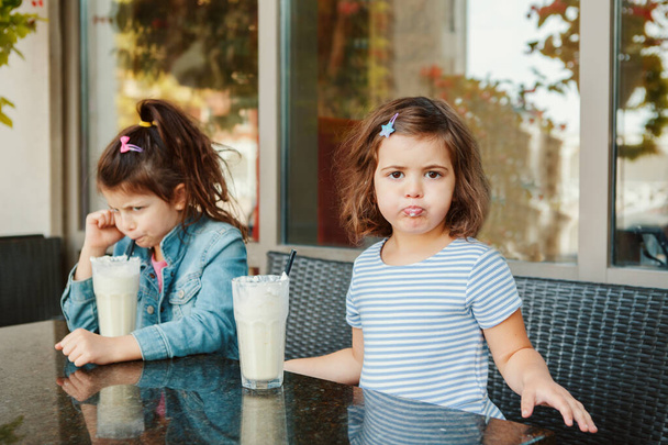Két vicces, mogorva, kaukázusi húgocska tejturmixot iszik a kávézóban. Barátnők veszekedtek. Hideg nyári desszertek gyerekeknek. Boldog autentikus gyermekkori életstílust.  - Fotó, kép