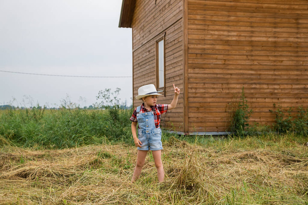 Aranyos kislány cowboy kalapban, farmernadrágban és piros fehér ingben, nyáron a faluban tölti az idejét, egy faház mellett áll. - Fotó, kép