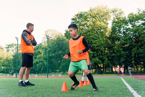 Concentrated junior soccer player running among orange cones that standing on artificial turf at stadium during workout - 写真・画像