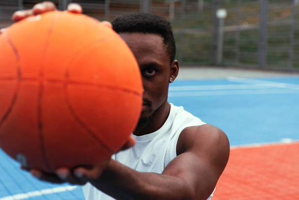 Waist portrait of a young African athlete with a joyful smile in a former T-shirt. Focus on the basketball that the guy is holding in front of him with his arm outstretched. - Photo, Image