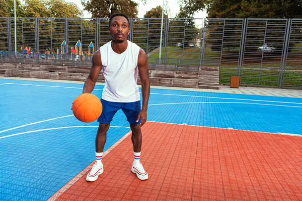 Basketbol sahasında beyaz tişörtlü ve mavi şortlu genç Afro-Amerikan basketbolcu. Basketbol sahasında, tasarlanmış, spor konsepti, sağlıklı hareket.. - Fotoğraf, Görsel