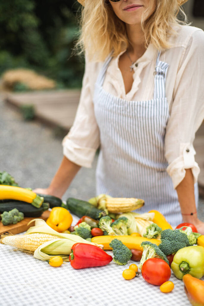 Die Sommerernte liegt auf dem Tisch. Viel Gemüse: Zucchini, Brokkoli, Tomaten, Mais, Kräuter, Paprika. Ein Bauernmädchen steht neben dem Tisch, auf dem Gemüse liegt. Hochwertiges Foto - Foto, Bild