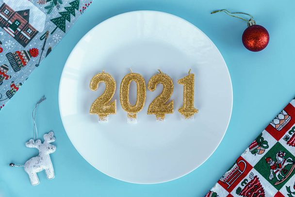 Xmas, winter, new year, festive table and treats, food concept - Blue Christmas background with white plate on which candles instead of dish in form of numbers 2021 with different accessories. Flatly - Foto, Imagen