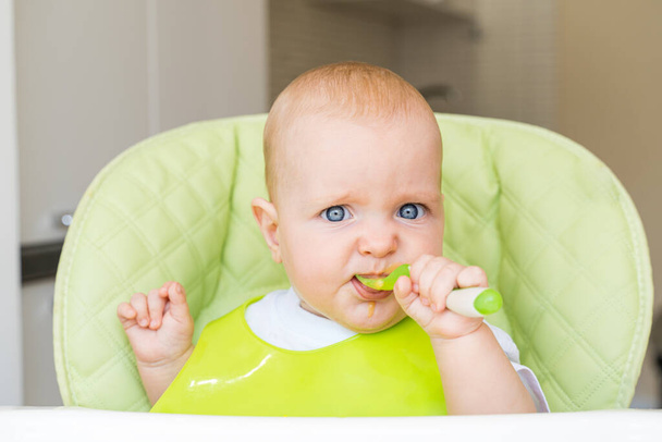 a small child with blue eyes and blond hair sits in a high chair and eats the first bait. Healthy diet. Baby 6 months - Foto, immagini