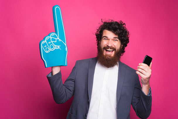 Portrait of cheerful bearded man in suit holding smartphone and pointing with fan glove - Φωτογραφία, εικόνα
