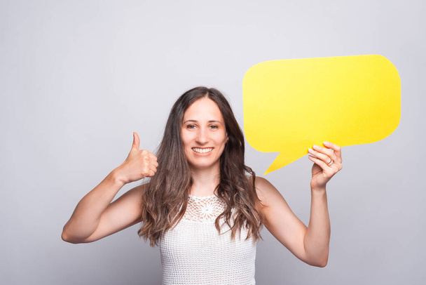 charming beautiful young woman showing thumb up gesture and holding empty blank speech bubble - Foto, immagini