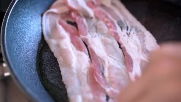 Close-up de tiras suculentas de bacon fritando na panela de cozinha com óleo, mãos de pessoas colocando fatias de bacon na superfície da panela quente e preparando delicioso café da manhã em casa. Fritura de bacon crocante no - Filmagem, Vídeo