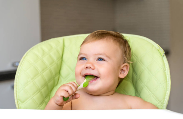 a small child with blue eyes and blond hair sits in a high chair and eats the first bait. Healthy diet. Baby 11 months - Φωτογραφία, εικόνα