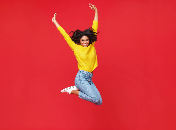 Πλήρες σώμα χαρούμενα έθνικ γυναίκα σε casual ρούχα χαμογελώντας και πηδώντας επάνω, ενώ διασκεδάζοντας κατά κόκκινο backdro - Φωτογραφία, εικόνα