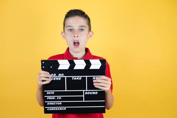 Young beautiful child boy over isolated yellow background surprised holding clapperboard - Фото, зображення