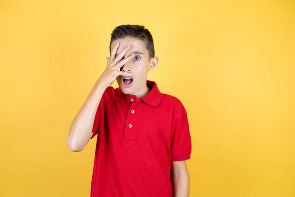 Young beautiful child boy over isolated yellow background peeking in shock covering face and eyes with hand, looking through fingers with embarrassed expression - Photo, Image