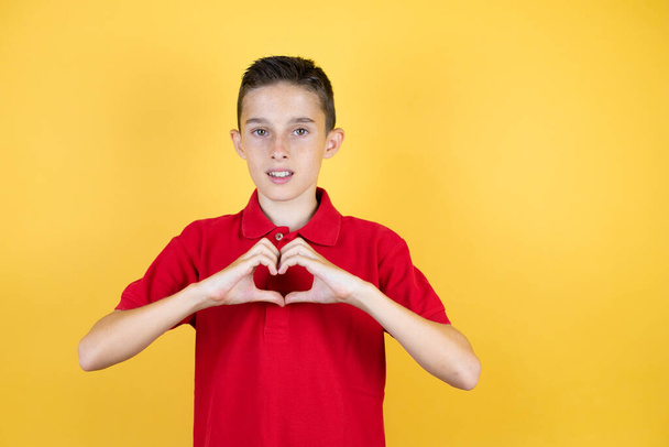 jong mooi kind jongen over geïsoleerde gele achtergrond glimlachen in liefde tonen hart symbool en vorm met handen - Foto, afbeelding