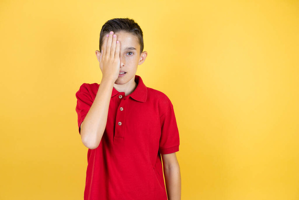 Young beautiful child boy over isolated yellow background covering one eye with hand, confident smile on face and surprise emotion. - Photo, Image