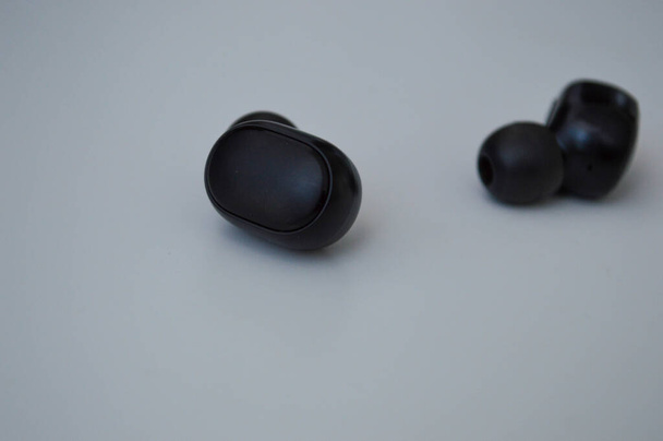 黒い小さなワイヤレスヘッドフォンは白い表面にあります。マクロ・アングルで音楽を聴くための装置です。美しいアクセサリー。便利な音楽切り替えボタン - 写真・画像