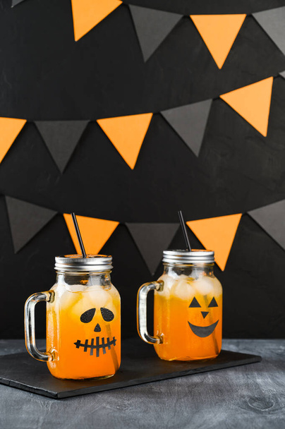 Halloween pumpkin iced mocktails in glass jars decorated with scary faces on a dark table. DIY Halloween Party decoration on black wall. - Foto, Imagen