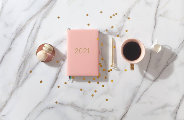 Ροζ κοραλλί ημερολόγιο για το έτος 2021, στυλό, καφέ latte, μπισκότο macaron και ψάθινο πλεκτό placemat σε λευκό μαρμάρινο φόντο. Πρωτοχρονιάτικο σχέδιο. Μινιμαλιστική θέση εργασίας. Αντιγραφή χώρου. - Φωτογραφία, εικόνα