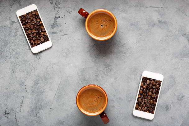 Zwei Kaffeetassen mit frischem und aromatischem Kaffee auf dem Tisch. Spritzer Kaffeebohnen im Fenster des Smartphones. Ansicht von oben. - Foto, Bild