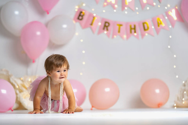First year's birthday.a happy little girl in a pink tutu skirt crawls on a background with garlands and pink balloons, celebrating her first birthday. Birthday decoration. Copy space. - Foto, Bild