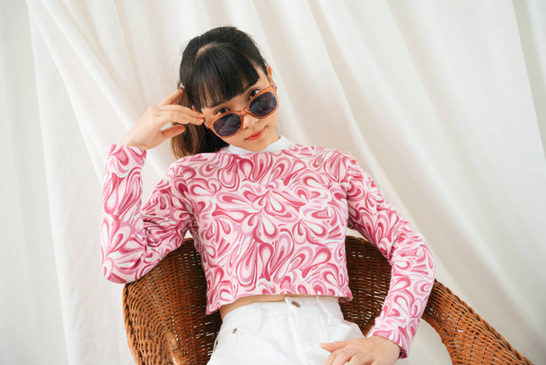 Divat portré fiatal ázsiai nő rózsaszín ing visel napszemüveg ül egy széken. - Fotó, kép