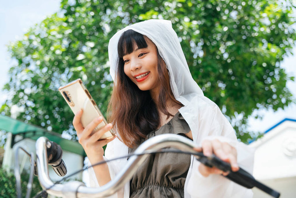 Νεαρή Ασιάτισσα φοράει λευκό αδιάβροχο χρησιμοποιώντας smartphone στο ποδήλατο στο πάρκο. - Φωτογραφία, εικόνα