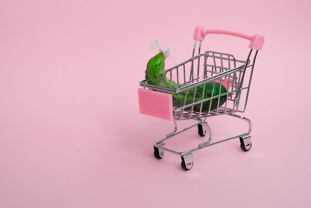 Pequeño carrito de comestibles con gran pepino inusual sobre fondo rosa - Foto, imagen