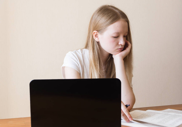 Девочка учится перед ноутбуком. Подросток с длинными светлыми волосами листает книгу. - Фото, изображение