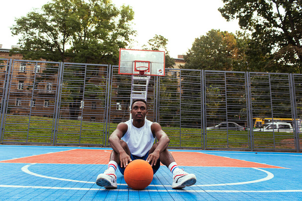 Cansado, o homem afro-americano senta-se no chão com a bola no campo ao ar livre. Faz uma pausa.. - Foto, Imagem