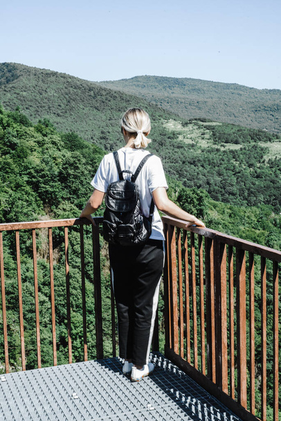 joven adolescente se para con su espalda con una mochila en la plataforma de observación con vistas a las montañas  - Foto, imagen