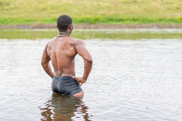 Młody facet o wysportowanej budowie stoi w pasie w wodzie jeziora plecami do kamery i patrzy w oddali. - Zdjęcie, obraz