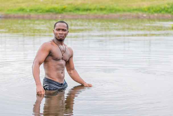 Een jonge man van atletische bouw staat taille-diep in het water van het meer en kijkt naar de camera - Foto, afbeelding