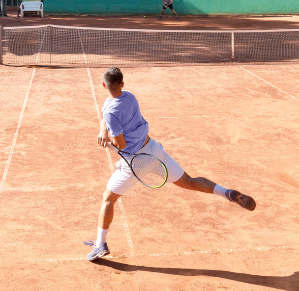 Young tennis player on clay tennis court with racket plays forehand hit. Professional tennis player in dynamic motion after hitting the ball. Sports action frame. Back view, shadow, square size - Photo, Image