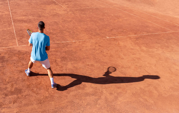 テニスの選手と赤い粘土のテニスコートで彼の影。プロの男性テニス選手はボールを受け取る準備ができています。テニスの試合開始。スポーツの背景。テキストのコピースペース - 写真・画像