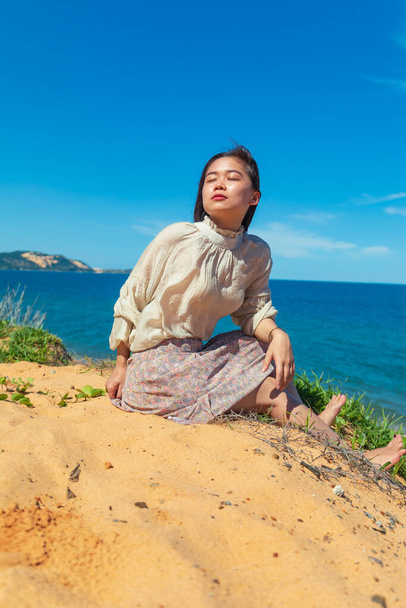 A vertical shot of a young attractive Asian female model posing while sitting on the sand at the beach - Fotó, kép