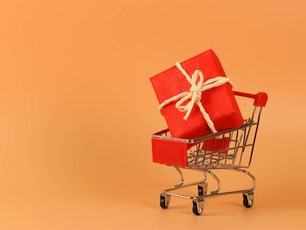 Vooraanzicht van rode geschenkdoos op winkelwagentje op oranje achtergrond met kopieerruimte. Kerstvakantie concept. - Foto, afbeelding