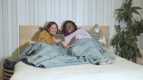 Afroamericani e donne europee sono sdraiati sul letto a guardare il social network, atmosfera familiare, accogliente appartamento la sera, Ulybayutsya ridere per leggere lo studio - Filmati, video