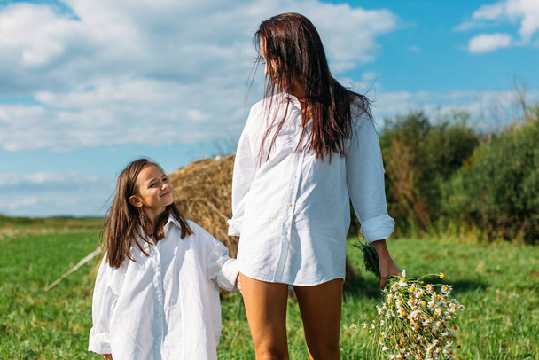 Mom and daughter, dressed alike, in white shirts and with flowers, walk in the fields in summer. around the sun, greenery, field, grass, hay, sky. - Photo, image