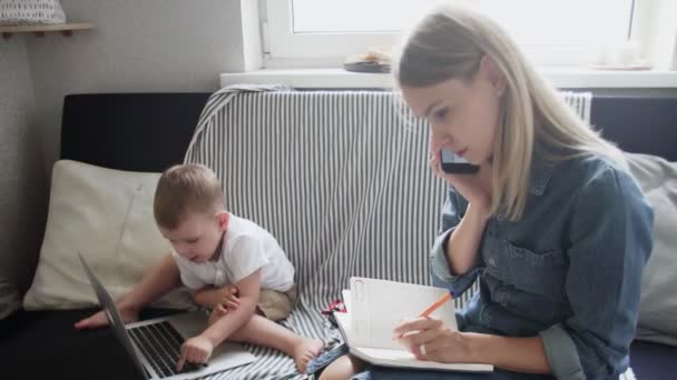 Mujer ocupada hablando por teléfono y tomando notas mientras su hijo juega con el cuaderno - Imágenes, Vídeo
