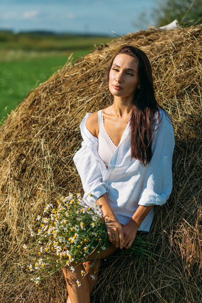 A young beautiful girl with long dark hair and a good figure, a girl in light underwear and a shirt, she walks in the field, summer, sun sky, sunset green fields, hay. - Photo, image
