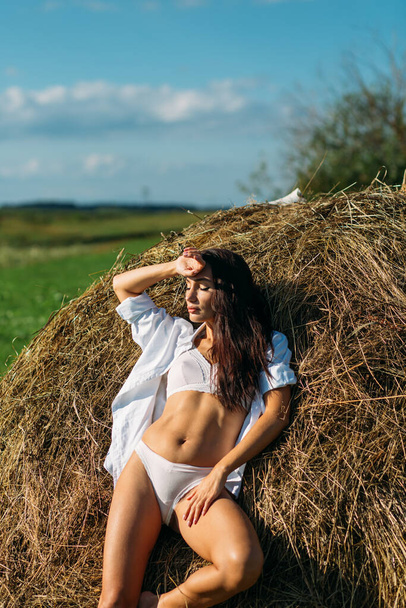 A young beautiful girl with long dark hair and a good figure, a girl in light underwear and a shirt, she walks in the field, summer, sun sky, sunset green fields, hay. - Фото, изображение