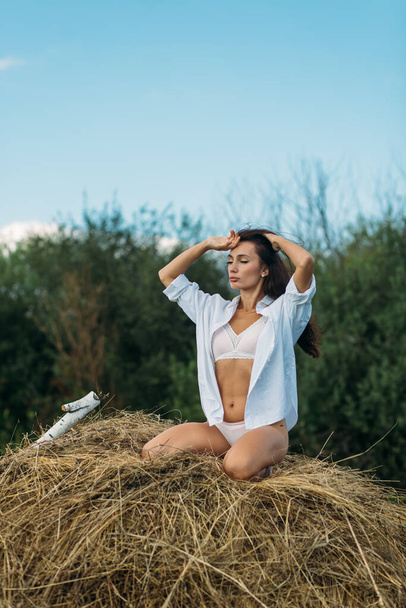 A young beautiful girl with long dark hair and a good figure, a girl in light underwear and a shirt, she walks in the field, summer, sun sky, sunset green fields, hay. - 写真・画像