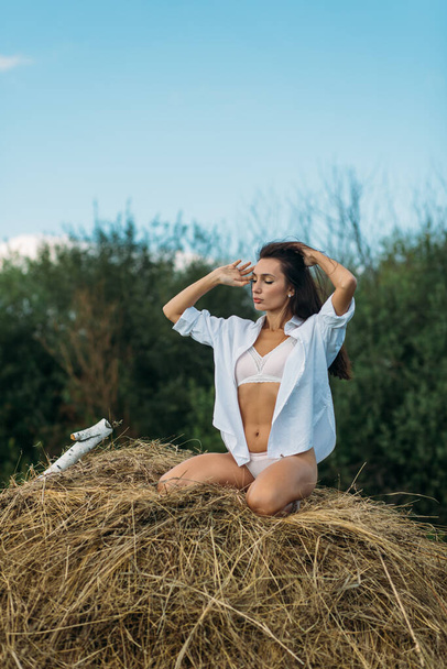 A young beautiful girl with long dark hair and a good figure, a girl in light underwear and a shirt, she walks in the field, summer, sun sky, sunset green fields, hay. - Foto, afbeelding