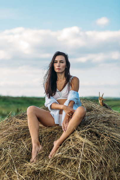 A young beautiful girl with long dark hair and a good figure, a girl in light underwear and a shirt, she walks in the field, summer, sun sky, sunset green fields, hay. - Фото, изображение