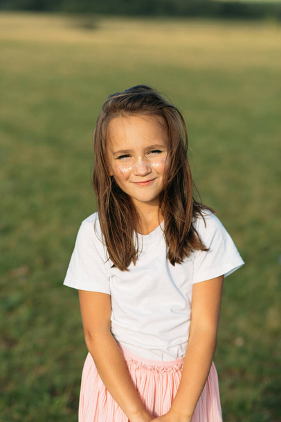 A little girl of about seven, with her hair down and a sunny shine on her cheeks. portrait. she stands in the middle of the field her face illuminates the evening sun, the girl is very happy - Photo, Image