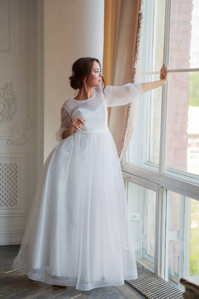 Full-length portrait of the bride in a white wedding dress by the window. - Zdjęcie, obraz