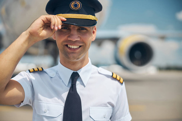 Όμορφος πιλότος στη διοίκηση αγγίζει καπέλο καπετάνιου και χαμογελά - Φωτογραφία, εικόνα