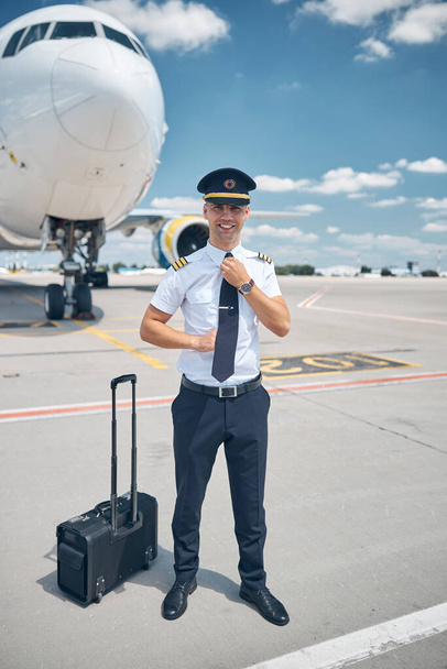 Όμορφος άντρας πιλότος προσαρμόζει τη γραβάτα του στο αεροδρόμιο. - Φωτογραφία, εικόνα