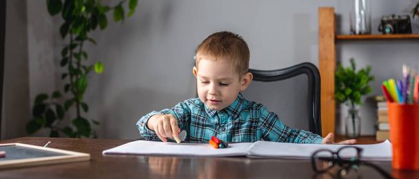 Милый мальчик рисует мелом на бумаге в альбоме за столом. Концепция дошкольного образования и развития творчества - Фото, изображение