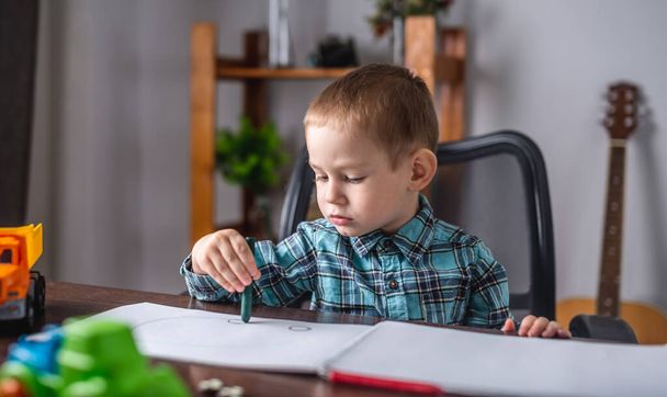 Χαριτωμένο παιδί ζωγραφίζει με κιμωλία σε ένα άλμπουμ στο τραπέζι. Έννοια της προσχολικής εκπαίδευσης και ανάπτυξη της δημιουργικότητας - Φωτογραφία, εικόνα