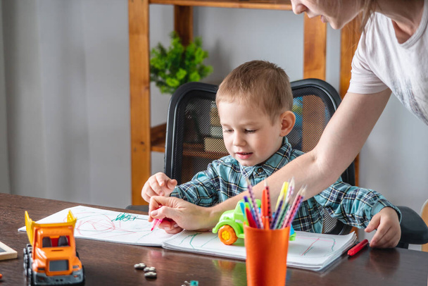 Η μαμά βοηθάει ένα χαριτωμένο παιδί να ζωγραφίσει με μολύβια σε ένα άλμπουμ στο τραπέζι. Έννοια της προσχολικής εκπαίδευσης και ανάπτυξη της δημιουργικότητας - Φωτογραφία, εικόνα